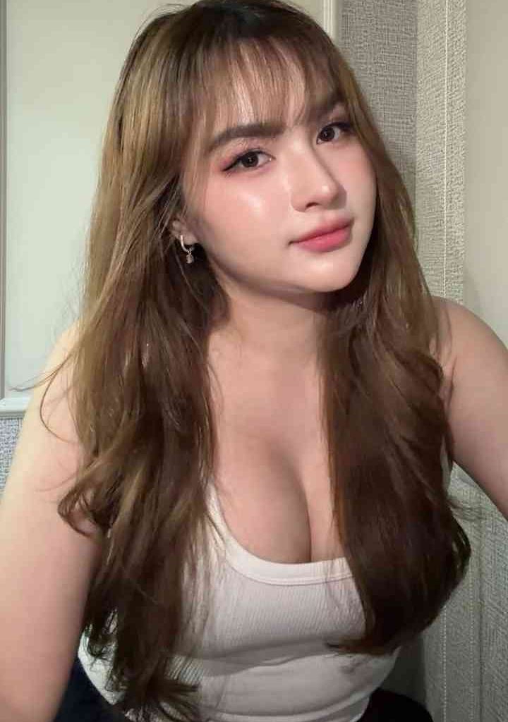 KL Escort Girl Young woman with long brown hair and bangs, wearing a white tank top. Escort Malay